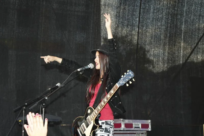 SuePrise mit Hut und E-Gitarre auf einer B&uuml;hne, eine Hand jubelnd erhoben, Publikum im Vordergrund